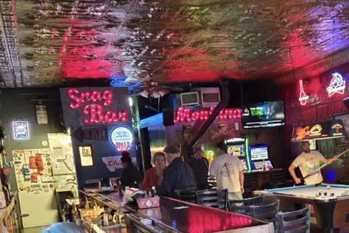The bar at the Snag, with bottles along the counter, the red neon Snag Bar Montana script on the wall behind it and the pressed tin ceiling overhead.