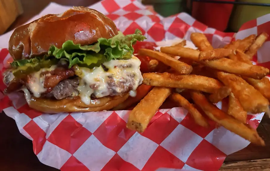 A bacon cheeseburger on a toasted pretzel bun, melted cheese running over the patty, with lettuce and pickles, sitting in a red and white checkered paper basket beside a pile of fries.