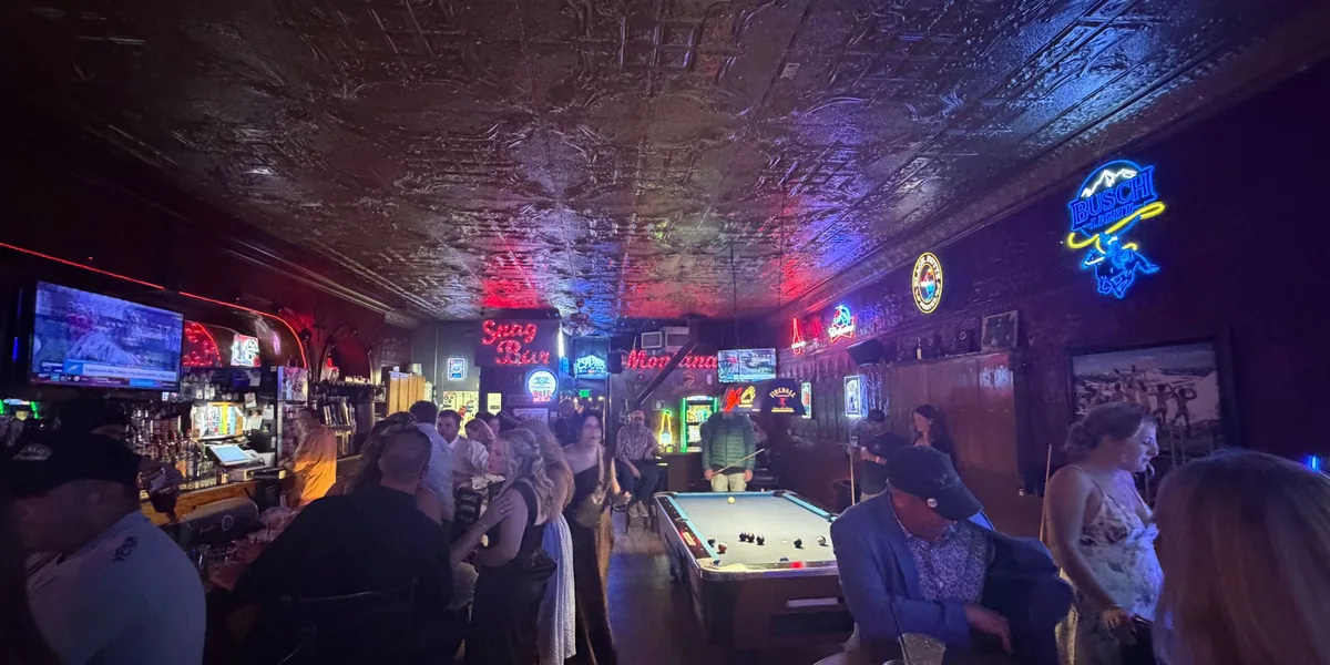 Inside the Snag Bar on a busy night: the pressed tin ceiling lit red and purple by the neon, the red Snag Bar and Montana neon signs above the back bar, a Busch sign on the wall, and a full room standing around the pool table.
