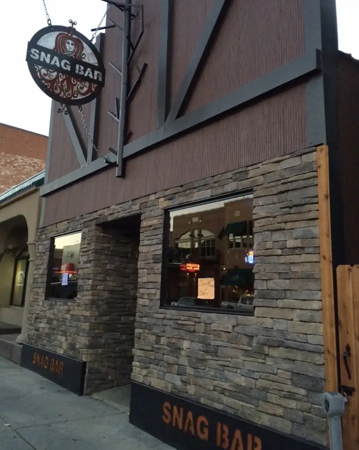 The Snag Bar on Broadway at dusk: a hanging oval sign carrying the bar&rsquo;s woman&rsquo;s-face artwork, a board-and-batten front above stacked stone, and SNAG BAR lettered in orange along the base.